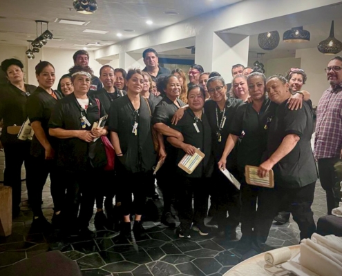 A group of two dozen Hotel Figueroa workers in black work uniforms pose emotionally in a tiled dining area.